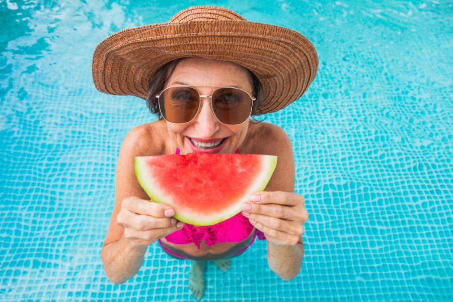 Woman standing in a swimming pool practicing UV safety with a wide brimmed hat, sunglasses, eating watermelon to stay hydrated